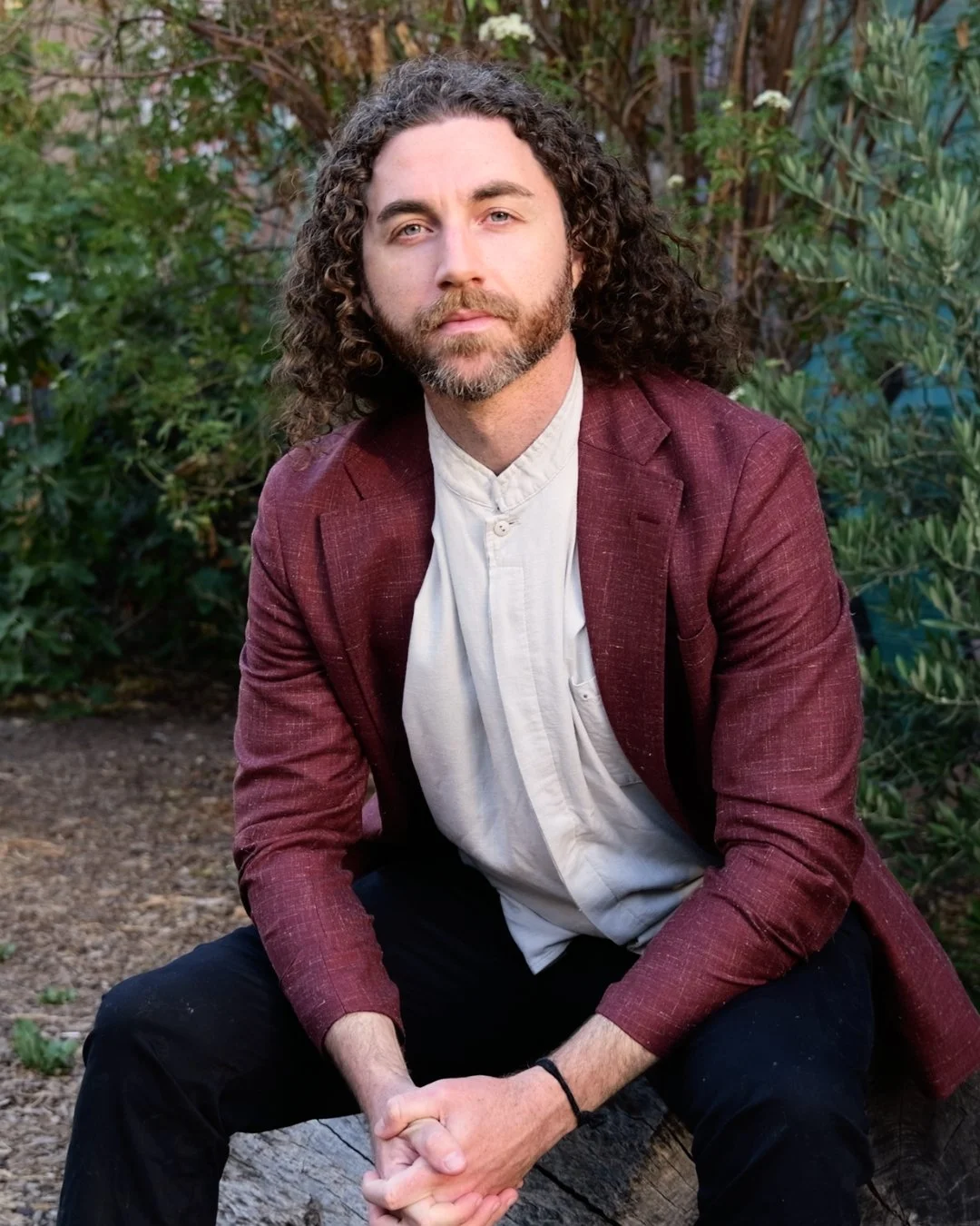 Tim Adalin in a burgundy blazer, seated outdoors in a green garden, leaning forward with hands clasped.