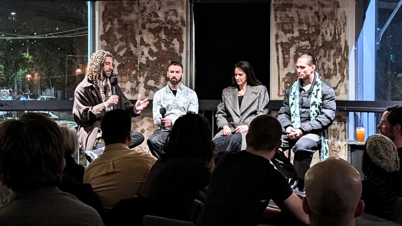 Tim Adalin, holding a microphone, hosting three panellists on a low stage, the audience in the foreground under warm light.