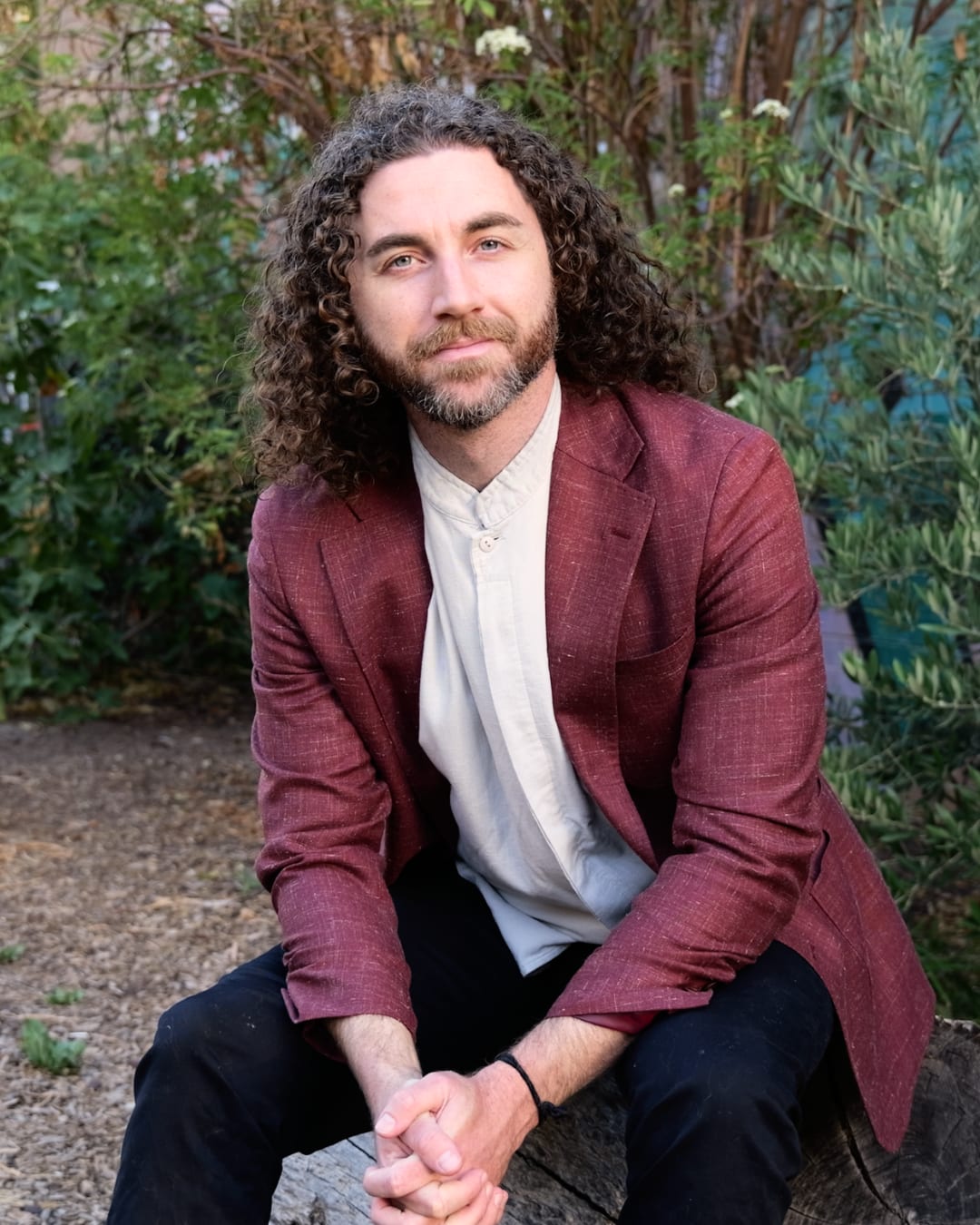 Tim Adalin, smiling slightly, seated outdoors in a burgundy blazer and white collarless shirt.