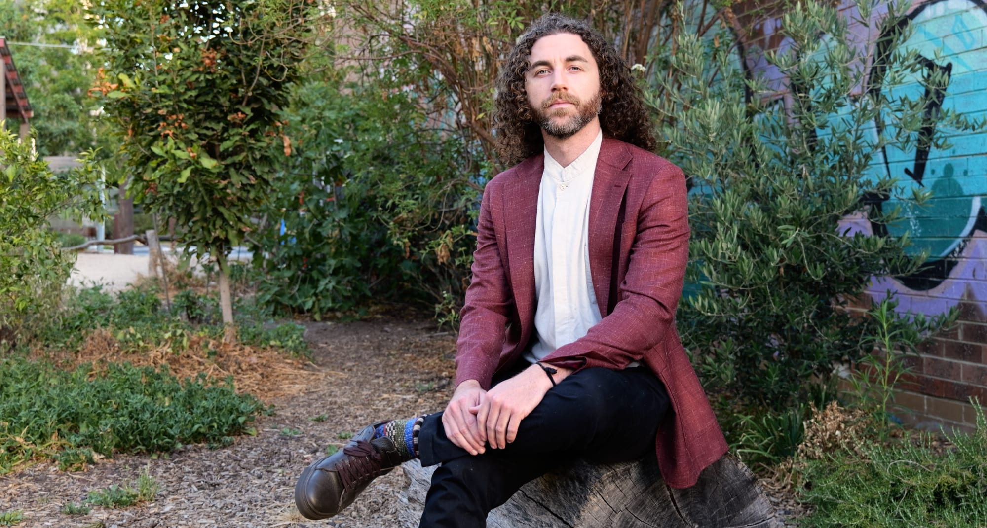 Tim Adalin in a burgundy blazer and white collarless shirt, seated on a log in a green urban garden, a teal and lavender mural behind him.