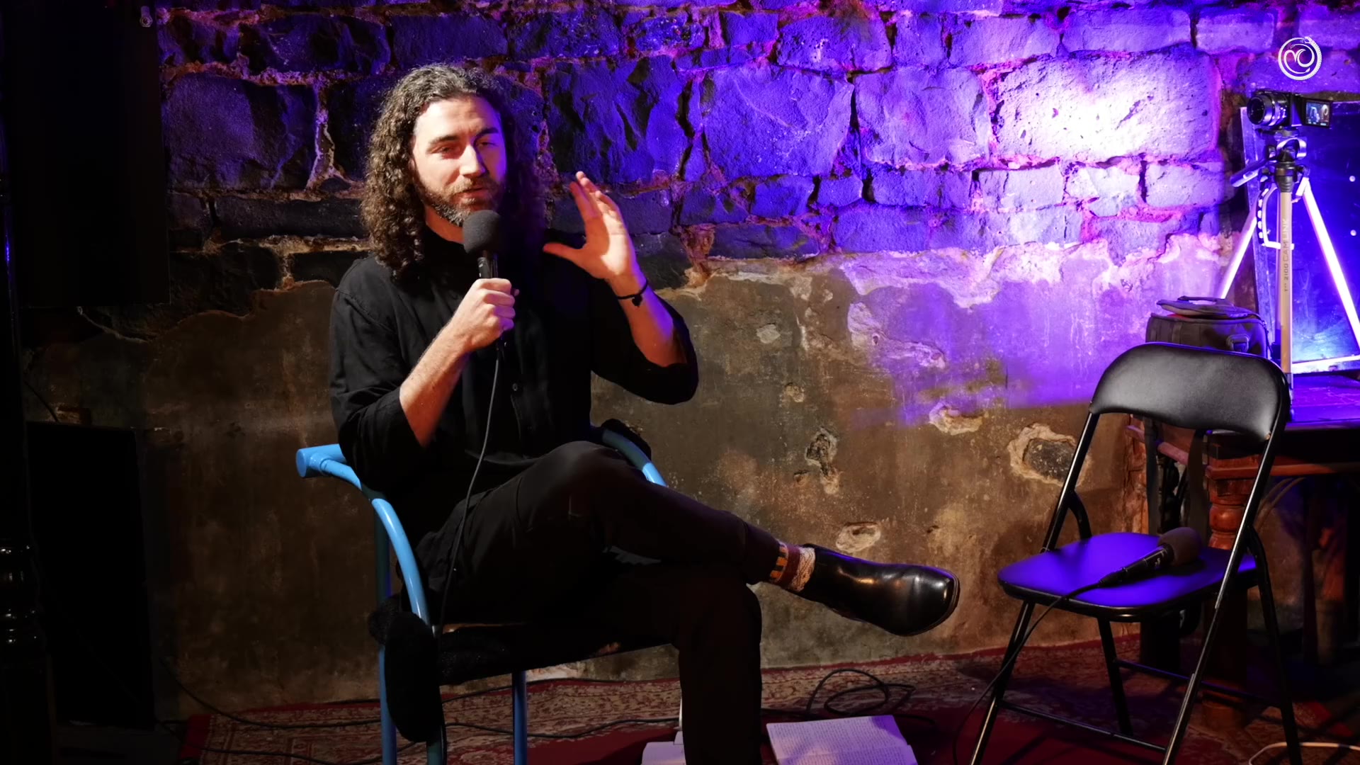 Tim Adalin seated on a blue chair with a microphone, one hand raised mid-thought, in a stone vault washed in violet light, at Underground Philosophy No. 14, Science and Magic.
