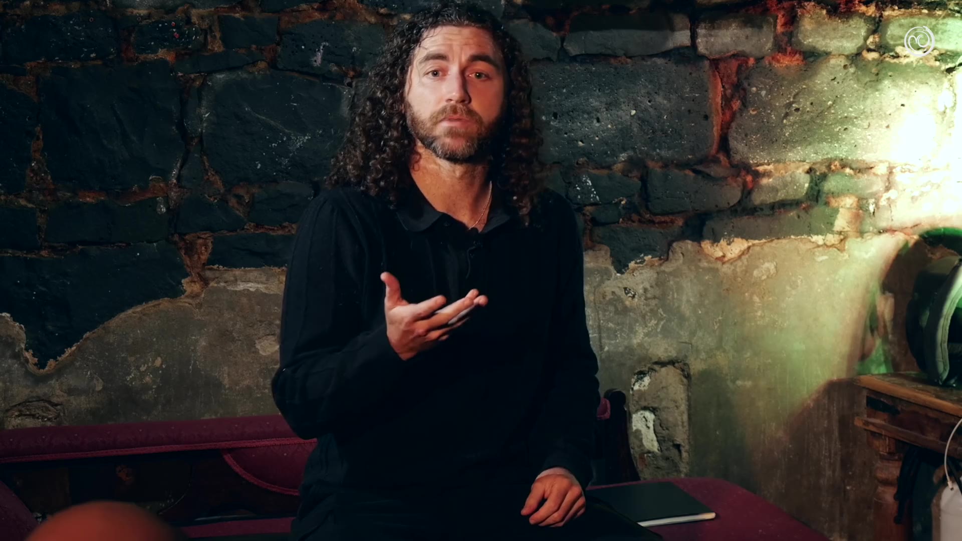 Tim Adalin in black, seated against a rough bluestone wall lit green from one side, speaking with one hand raised, at Underground Philosophy.