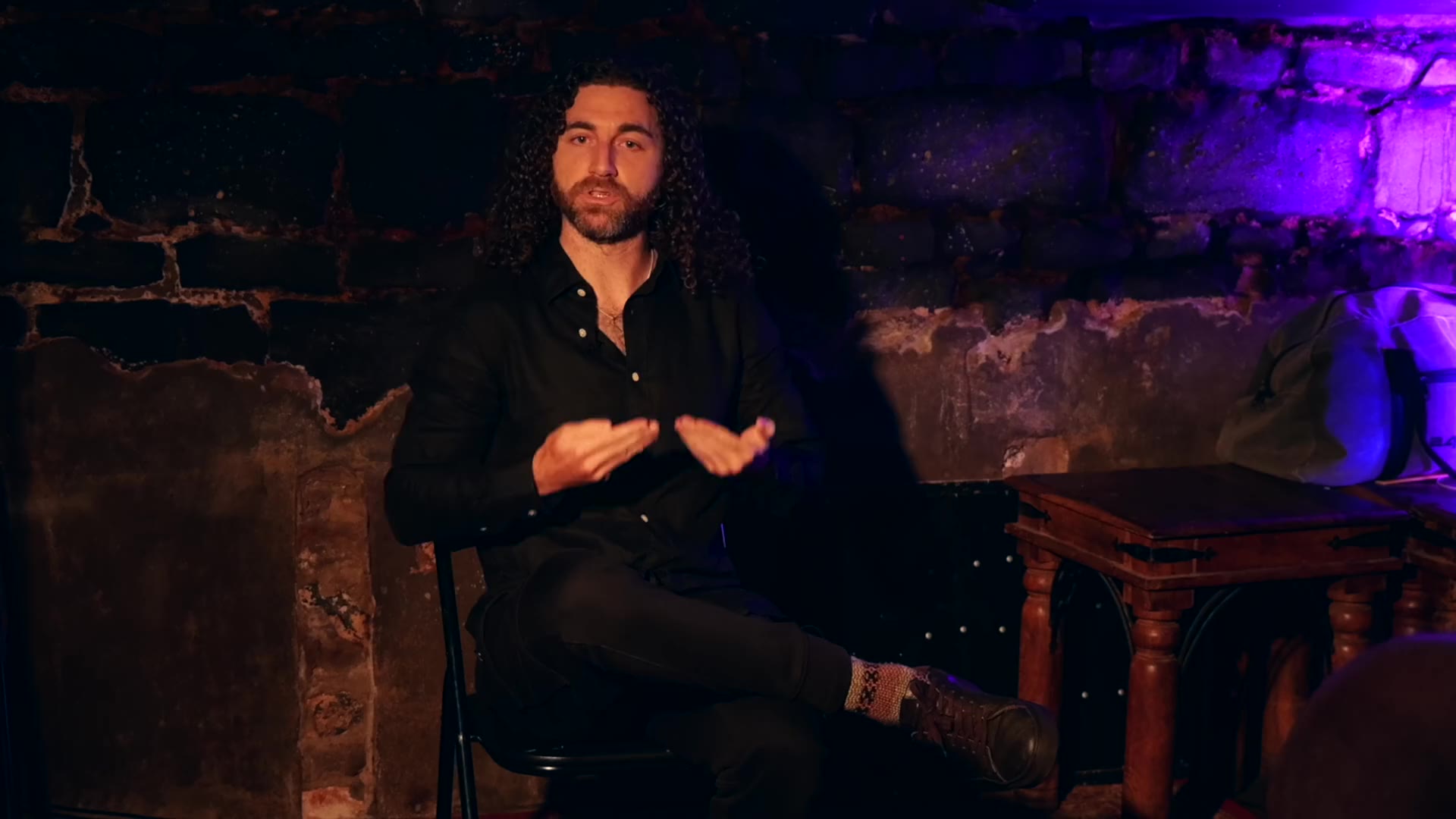Tim Adalin in a black shirt, seated, speaking with open hands in a darkened vault lit with violet, at Underground Philosophy No. 11, Truth to Power.